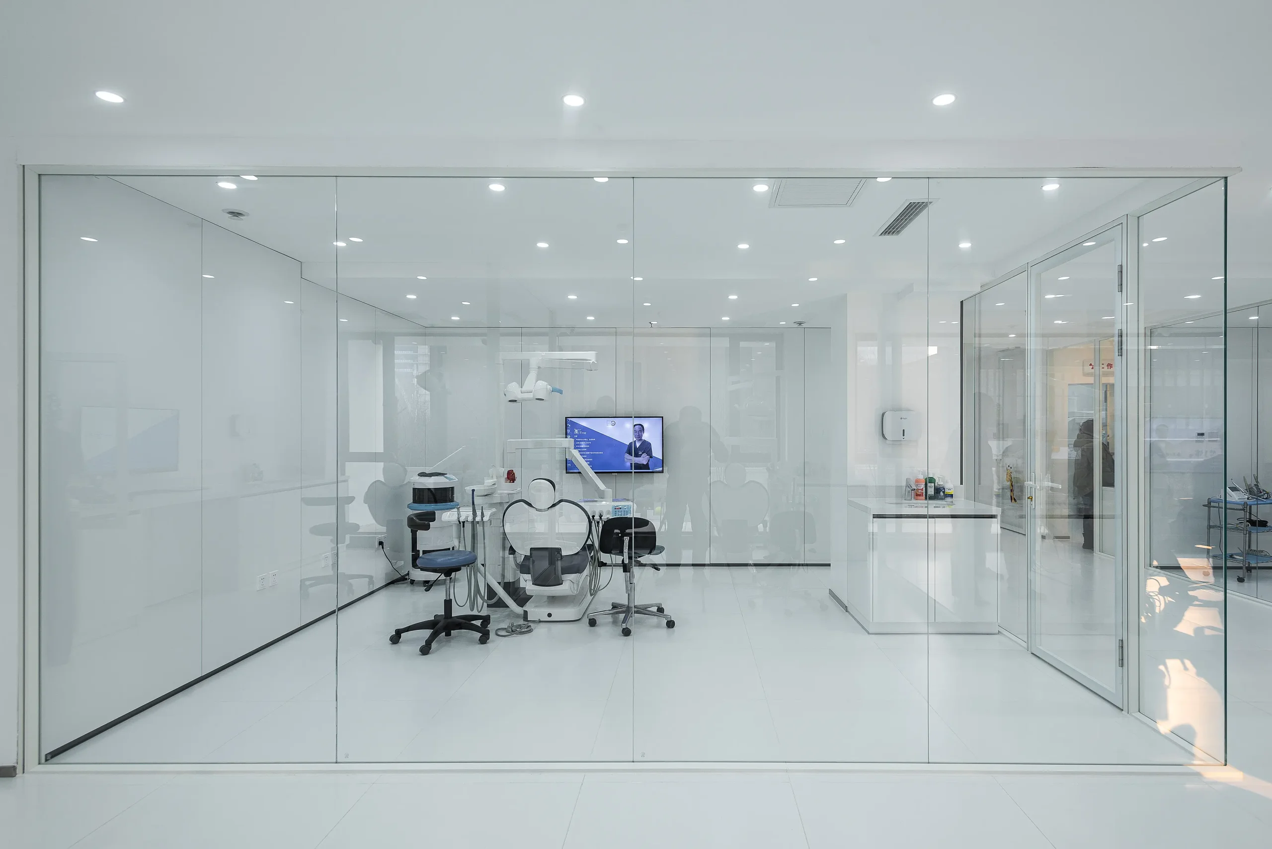 Dental clinic partition enclosing a single operatory in frameless glass, with the treatment chair and delivery unit visible from the corridor